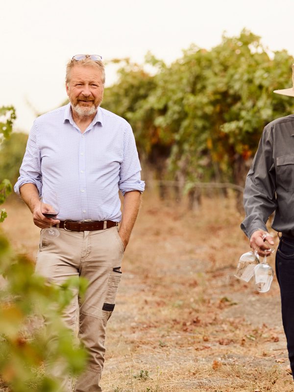 Two men walking through vineyard rows holding wine glasses and a bottle of Brecon Estate wine.