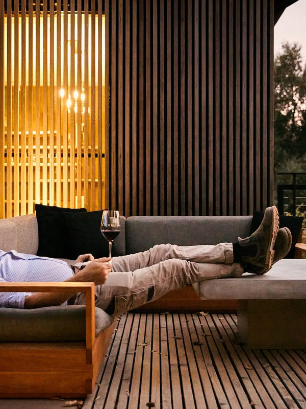 Man lounging on an outdoor patio sofa with a wine glass outside a modern illuminated building.