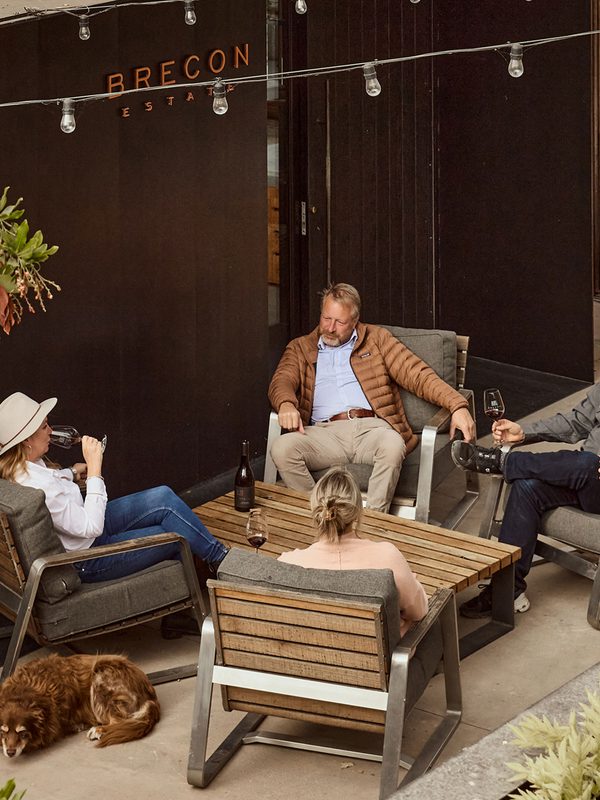 Group sitting at an outdoor patio table enjoying wine outside the modern Brecon Estate building with a dog lounging nearby.