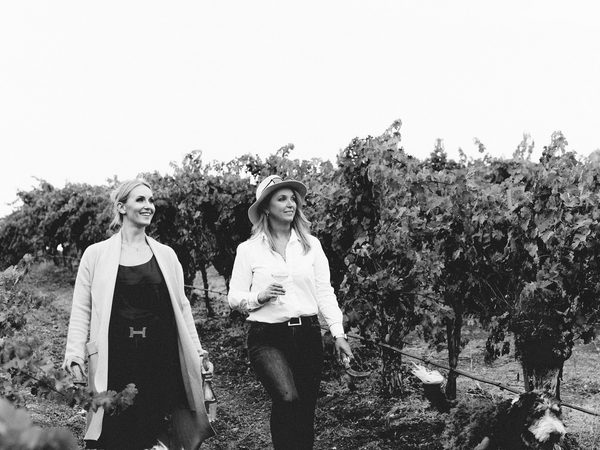 Black and white photo of two women walking through a vineyard holding wine glasses while dogs walk alongside them.