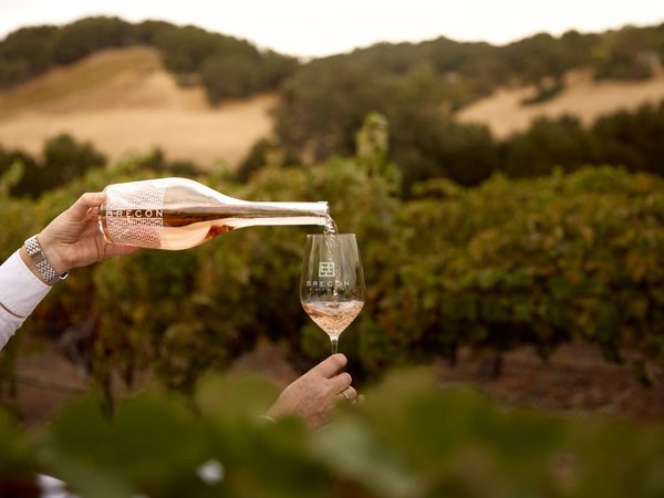 Rosé wine being poured into a branded Brecon Estate glass outdoors in front of a vineyard and rolling hills.